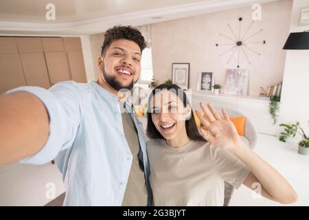 Young woman waving hand while her husband making selfie in living-room Stock Photo