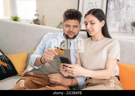 Young man with credit card and his wife with smartphone choosing goods in online shop Stock Photo