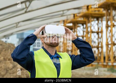Serious builder in uniform and vr headset standing on construction site Stock Photo