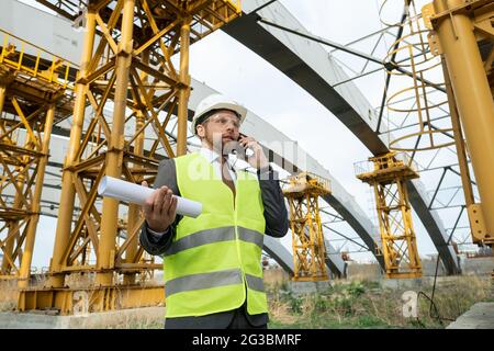 Young engineer in uniform holding smartphone and rolled blueprint Stock Photo
