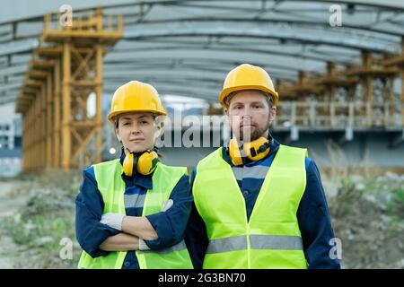 Two serious construction workers in uniform and helmets standing next to one another Stock Photo