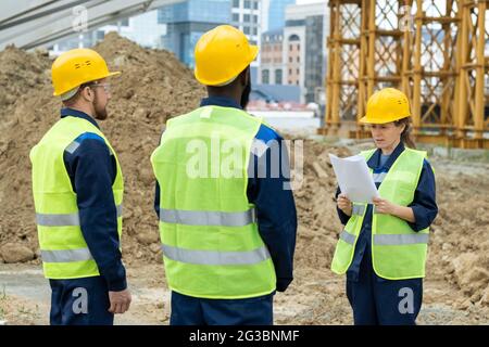 Two intercultural male builders listening to their female colleague with paper at working meeting Stock Photo