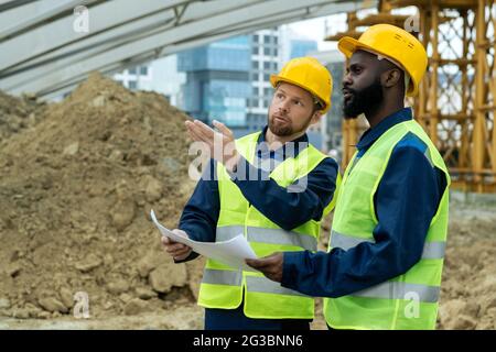 Two young intercultural builders in uniform discussing sketch and new construction Stock Photo
