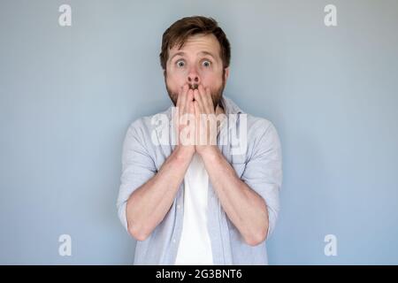 Shocked man in casual clothes puts hands over his mouth in fear and looks at the camera with big eyes. Gray background. Stock Photo
