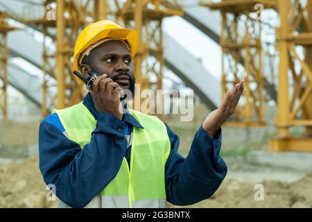 African foreman in workwear speaking on walkie-talkie at construction site Stock Photo