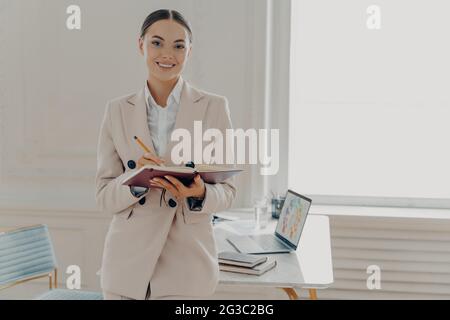 Positive happy female director in elegant wear planing working day while standing in office Stock Photo