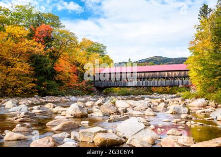 Old covered bridge spanning a mountain river with forested banks on a partly cloudy autumn day. Stunning autumn colours.