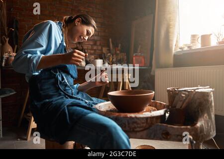 Beautiful young Caucasian female artisan wearing apron working with non painted clay pottery tableware in workshop Stock Photo