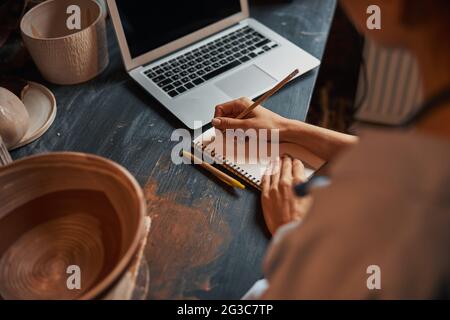 Unrecognized female artisan sitting at the work desk and looking to the screen of her laptop in pottery workshop Stock Photo