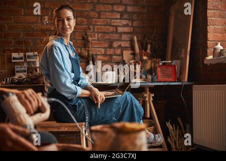 Brunette Caucasian craft woman sitting on the chair while posing at the photo camera in pottery workshop Stock Photo