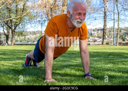 Portrait of a senior man exercising, doing push ups in the park. An elderly man in good shape with strong arms. Stock Photo