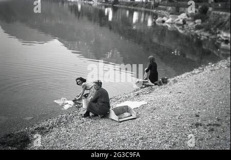 1950, two women on a shore by a lake hand washing garments using a form of washboard, a wooden washing box and board, France. This three-sided wooden box or caisse had a back half with a place to kneel on and front half (or washboard) where the clothes would be scrubbed or washed with the water. The woman knelt in the box and her skirt stayed dry. For more comfort, it would be padded with straw or a cushion as seen here. In some areas of the world, doing the laundry by a local river or lake, a tiring yet effective method, is still practised today. Stock Photo