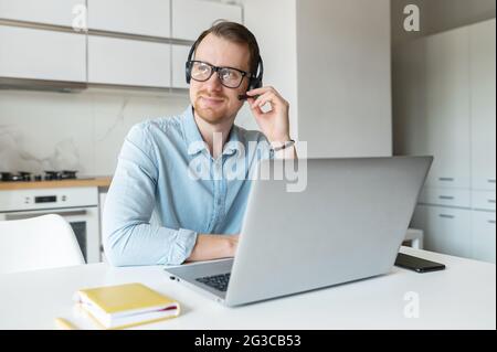 Smiling young male freelancer wearing a headset and looking away, a positive man with the beard working remotely in the customer service department, making and receiving calls Stock Photo