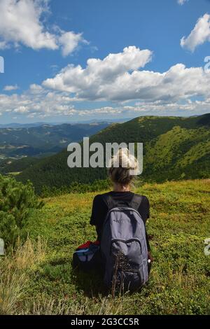 Girl sitting with her back on top of a mountain in the summer with a backpack. Stock Photo