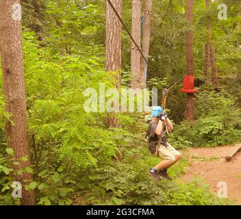 Boy on a sliding swing. Child in forest adventure park. Kids climb on high rope trail. Kid in a blue helmet on a sliding swing. Agility skills and Stock Photo