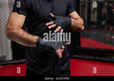 cropped man in black sportswear preparing for tough fight, wrapping fist in sport protective bandages. standing on ring getting ready for mma fight Stock Photo