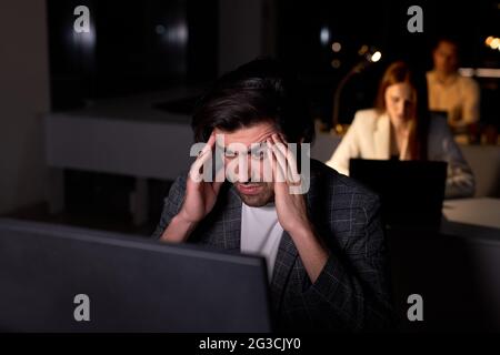 Portrait of tired male in suit sitting at desk in office using computer pc, thinking, having hard task, no time to sleep. side view on brunette guy of Stock Photo
