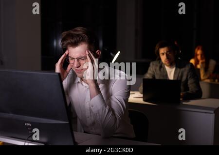 Young Caucasian Guy In Formal Wear Having Headache And Chronic Work Fatigue, Need More Sleep And Rest. Exhausted Man Sitting At Desk Using Pc Computer Stock Photo