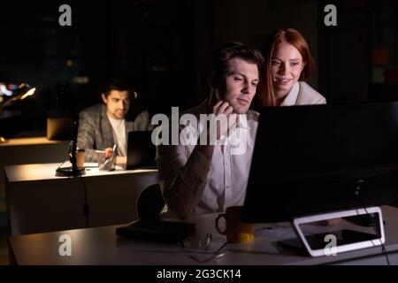 Two pleasant business people working together on computer, discussing project in office at late night. side view on redhead lady and handsome guy coll Stock Photo