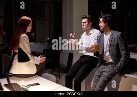 Side view on friendly office workers, colleagues having talk after hard working day, at night. Redhead woman and two guys in formal wear discussing, t Stock Photo
