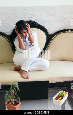 Full-length woman crying while sitting in an armchair near the laptop in the living room of her home. Stock Photo