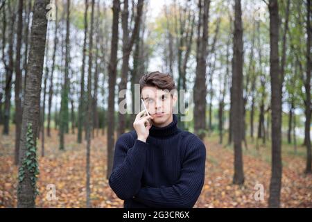 A man in a forest during autumn makes a phone call, all the leaves on the trees are yellow and many leaves have fallen to the ground. Stock Photo