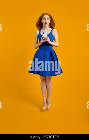 Full size profile photo of surprised redhead female in dress jump use smartphone search discounts run fast, isolated yellow color background in studio Stock Photo