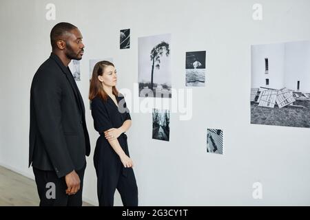 Portrait of elegant mixed-race couple wearing black while looking at photographs in modern art gallery, copy space Stock Photo