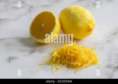 one full lemon, a second sliced and a pile of zest on a marble surface with natural light Stock Photo