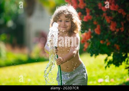 American child playing with garden hose in backyard. Funny excited kid having fun with spray of water on yard nature background. Summer kids outdoors Stock Photo