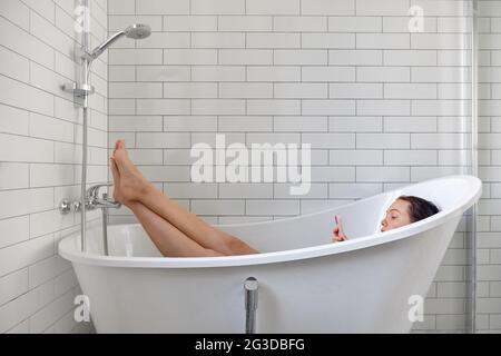 Young female browsing internet on mobile phone while taking bath and relaxing in bathtub Stock Photo