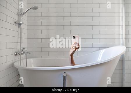 Anonymous female with modern pink mobile phone taking selfie while lying in bathtub and enjoying bath procedure in white bathroom