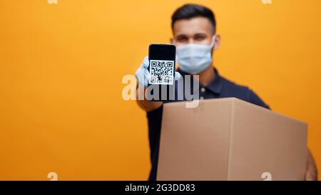 Man wearing a mask and gloves holding cardboard parcel box showing mobile phone with QR Code on the screen. Yellow background studio. Concept of E-wallet or Digital pay.  Stock Photo