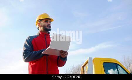 Young Worker wearing safety yellow helmet holding a laptop in his hand searching for address. A yellow truck can be seen behind him. Clear blue sky in the background. High quality daytime shot. Stock Photo