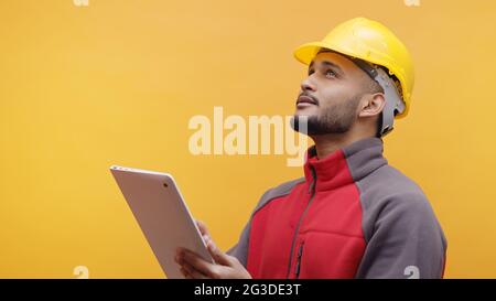 A young Engineer wearing yellow safety helmet holding a tablet in his hand. Studio shot with yellow background. Engineering and construction concept.  Stock Photo