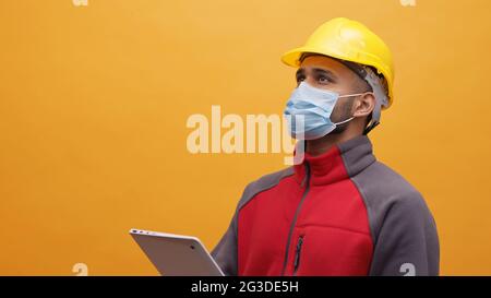 A young Engineer wearing yellow safety helmet and a mask holding a tablet in his hand. Studio shot with yellow background. Engineering and construction concept during Covid 19.  Stock Photo