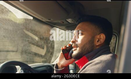 Young delivery guy driving a van while talking on phone. Guy holding phone in his hand. Parcel delivery concept. Sunlight reflecting his face. Daytime shot.  Stock Photo