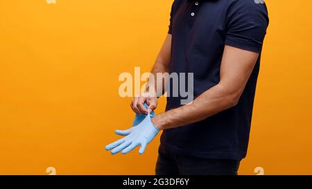 Delivery man putting on the gloves and taking precautions. Dressed in blue shirt. Face is not shown. Concept of Covid 19 and Pandemic. Yellow background studio shot.  Stock Photo