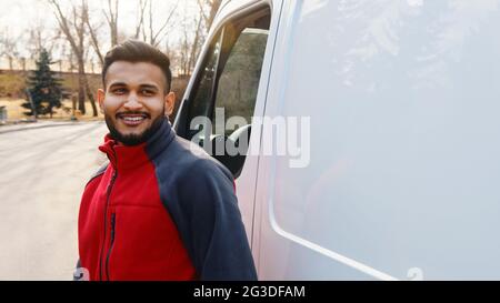Young Indian guy standing on the road with a white van behind him. Delivery guy wearing red uniform smiling. Trees in the background. Daytime shot.  Stock Photo