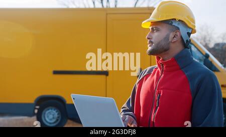 Delivery boy wearing a uniform and safety yellow helmet searching for address using a tablet standing with a yellow van behind him. Concept of parcel delivery service.  Stock Photo