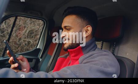 Young delivery guy sitting in the front seat of the van with hand on steering. Holding a phone in his other hand and smiling. Using his phone. Wearing a red uniform. Courier concept.  Stock Photo
