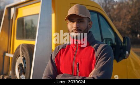Young worker standing with arms crossed and smiling. Dressed in red uniform and a cap. Yellow truck can be seen behind him with a spare tire. Concept of automobile mechanics. Closeup shot.  Stock Photo