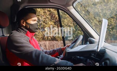 Young guy driving the car holding steering and gear. A laptop is placed on the dashboard. Dressed in red upper wearing a mask sitting on the driving seat of the car with a laptop in front of him.  Stock Photo