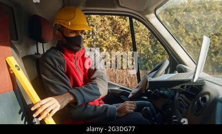 Man looking at the laptop placed on the dashboard of the car. Dressed in red upper wearing a mask and yellow helmet sitting on the driving seat of the car with a laptop in front of him. Stock Photo