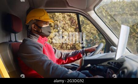 Man wearing yellow helmet and a mask driving a van. Focused man with hands on steering wheel looking straight ahead. Dressed in red upper. Trees in the background can be seen on a bright sunny day.  Stock Photo