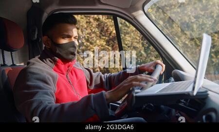 Indian man looking at the laptop placed on the dashboard of the car. Dressed in red upper wearing a black mask sitting on the driving seat of the car using a laptop placed on the dashboard of the car. Stock Photo