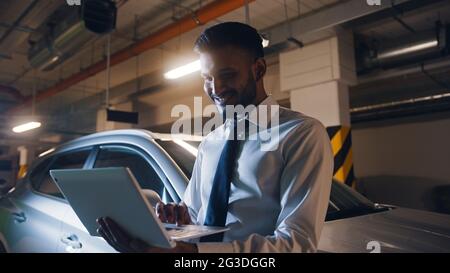 Businessman wearing formal shirt and tie in the parking lot with a laptop in his hand, smiling and using laptop. A white car is parked behind him.  Stock Photo