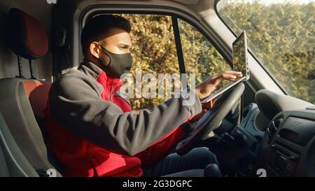 Indian guy sitting on the driving seat of the van using a laptop. Dressed in red zipper wearing a black Mask. Concept of car repair and maintenance. Sunlight hitting his face.  Stock Photo