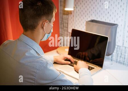 a young Caucasian man in a light shirt sits at a table in the evening at home wearing a protective medical mask and a mask and works at a laptop, remote work at home online Stock Photo