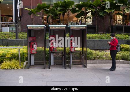 14.06.2021, Singapore, Republic of Singapore, Asia - A woman stands next to public telephone booths on Orchard Road browsing her mobile phone. Stock Photo